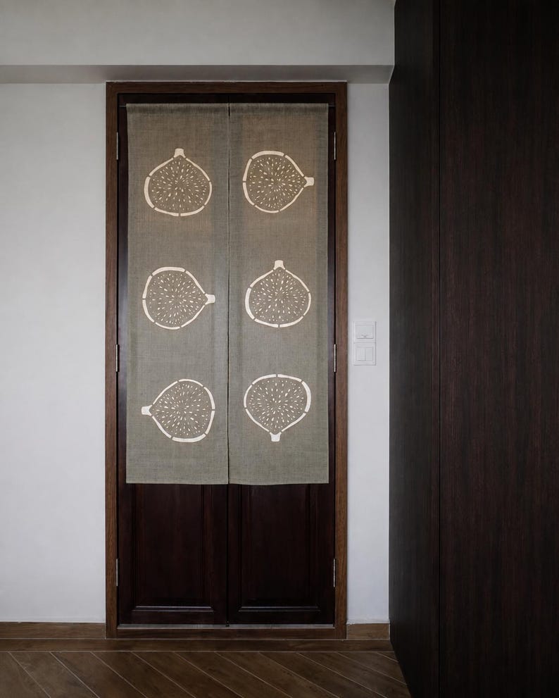 Puede incluir: Una cortina de puerta rectangular de tela beige con un estampado gr&aacute;fico blanco repetitivo de seis higos cortados por la mitad, dispuestos en dos columnas verticales. La cortina est&aacute; dividida en el centro y cuelga de un marco de puerta de madera oscura.