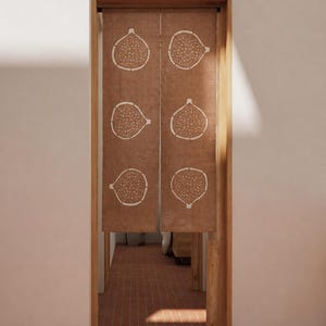 Puede incluir: Una cortina noren de tela marr&oacute;n colgada en una puerta, con un patr&oacute;n repetitivo de l&iacute;neas blancas de seis higos cortados por la mitad dispuestos en dos columnas verticales. El tejido tiene una textura visible y cuelga de un marco.