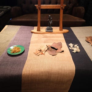 Puede incluir: Un camino de mesa de tela multicolor con rayas verticales en verde oliva, morado oscuro, beige y gris carb&oacute;n. El camino de mesa se muestra con platos peque&ntilde;os de hierbas secas, palitos de madera finos y varias piezas bot&aacute;nicas secas encima.