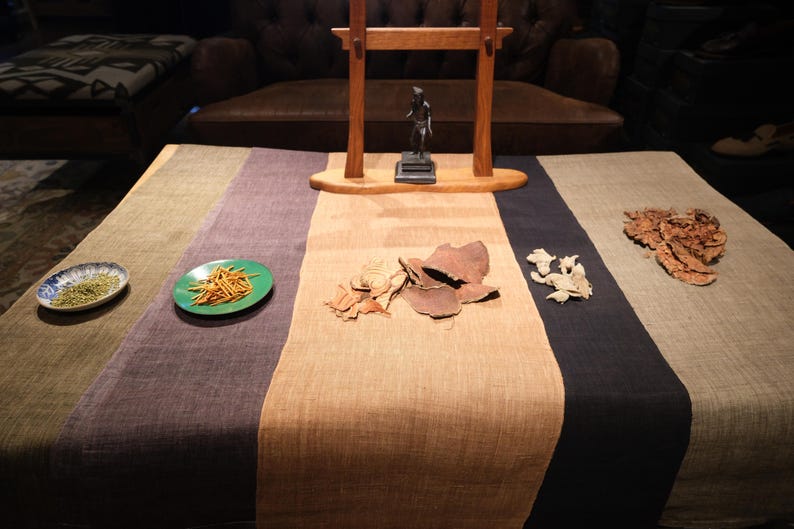 Puede incluir: Un camino de mesa de tela multicolor con rayas verticales en verde oliva, morado oscuro, beige y gris carb&oacute;n. El camino de mesa se muestra con platos peque&ntilde;os de hierbas secas, palitos de madera finos y varias piezas bot&aacute;nicas secas encima.