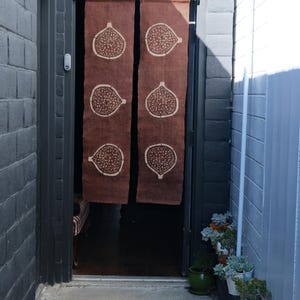 Puede incluir: Una cortina de puerta de tela marr&oacute;n de dos paneles con un patr&oacute;n repetitivo de higos en color crema. La cortina cuelga de una barra negra en el marco de una puerta, mostrando tres ilustraciones verticales de higos en cada panel.