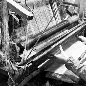 Puede incluir: Una vista en blanco y negro de un telar de madera tradicional con un gran cilindro que sostiene los hilos de urdimbre. El mecanismo cuenta con un pesado peine de madera y un rodillo de tela tensada para el proceso de tejido.