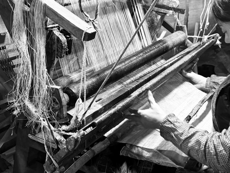 Puede incluir: Una vista en blanco y negro de un telar de madera tradicional con un gran cilindro que sostiene los hilos de urdimbre. El mecanismo cuenta con un pesado peine de madera y un rodillo de tela tensada para el proceso de tejido.