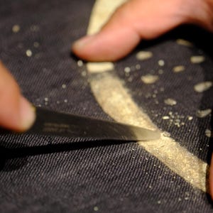 Puede incluir: Una hoja de metal afilada raspa a lo largo de una l&iacute;nea curva de color claro sobre una superficie de tela oscura y texturizada. Peque&ntilde;as part&iacute;culas de material claro est&aacute;n esparcidas a lo largo del camino de la hoja sobre el tejido negro.