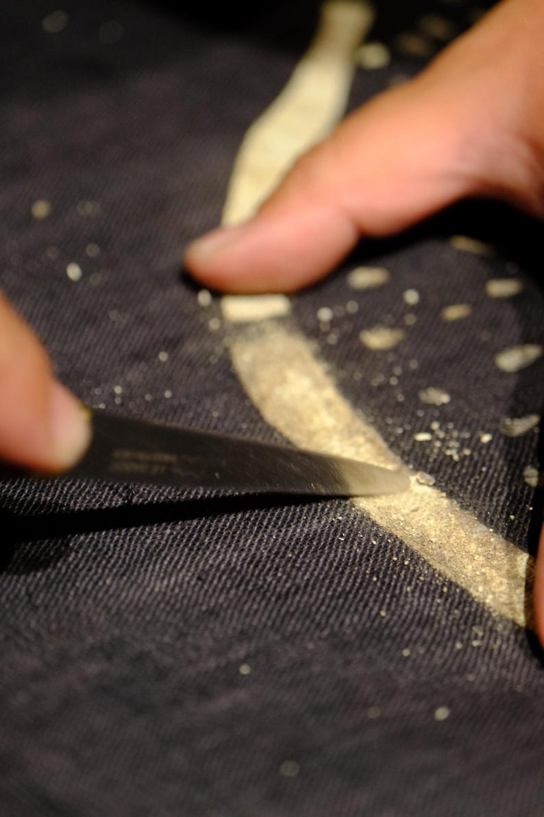 Puede incluir: Una hoja de metal afilada raspa a lo largo de una l&iacute;nea curva de color claro sobre una superficie de tela oscura y texturizada. Peque&ntilde;as part&iacute;culas de material claro est&aacute;n esparcidas a lo largo del camino de la hoja sobre el tejido negro.