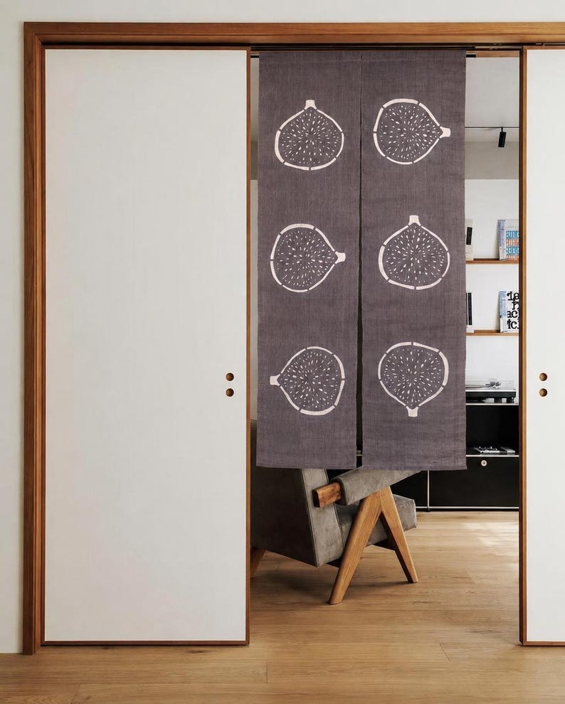 Puede incluir: Una cortina noren de tela gris oscuro con un patr&oacute;n repetitivo de l&iacute;neas blancas de higos cortados por la mitad. La cortina rectangular est&aacute; dividida en dos paneles verticales y cuelga en una puerta, mostrando seis ilustraciones de higos.