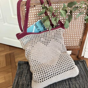Könnte beinhalten: Eine beige Häkeltasche mit einem herzförmigen Muster in der Mitte, akzentuiert durch einen dunkelroten Rand und zwei passende Schulterriemen. Die Tasche hat ein offenes Netzdesign und enthält ein Buch mit blau-weiß gestreiftem Einbandtext.