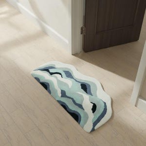 Puede incluir: Una alfombra semicircular con un diseño ondulado en tonos azules, turquesas y blancos. La alfombra está colocada sobre un suelo de madera clara cerca de una puerta. El patrón de la alfombra se asemeja a un paisaje estilizado.