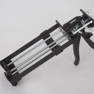 May include: A black dual-component caulk gun with a trigger and a handle. The gun has a metal frame with two parallel rods and a nozzle holder. The background is plain white.