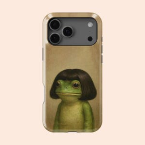 May include: A phone case featuring a green frog with a dark brown wig. The frog has large, expressive eyes and a neutral expression. The case has a tan background and a camera cutout.