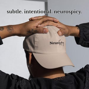 Puede incluir: Gorra de béisbol beige con la palabra "NeuroSpicy" en escritura negra. La gorra la sostiene una persona con tatuajes en el brazo. La imagen también incluye las palabras "subtle. intentional. neurospicy."