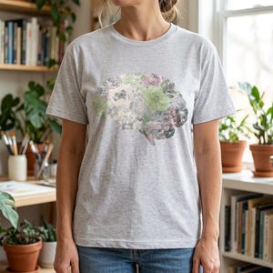 Op de afbeelding: Een lichtgrijs T-shirt met korte mouwen en een centrale grafische print van een menselijk brein, samengesteld uit diverse bloemen en vetplanten in gedempte tinten groen, paars en wit, gecombineerd met een blauwe spijkerbroek.