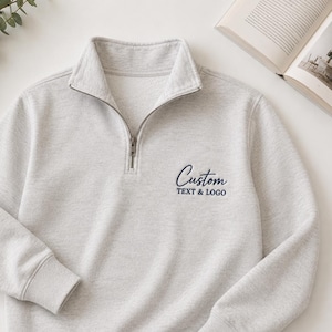 Könnte beinhalten: Ein hellgraues Sweatshirt mit einem Kragen mit Viertelreißverschluss. Das Sweatshirt hat lange Ärmel und eine gerippte Manschette. Der Text "Custom TEXT & LOGO" ist in Marineblau auf der Vorderseite gestickt. Ein Zweig Grün und ein offenes Buch sind im Hintergrund.