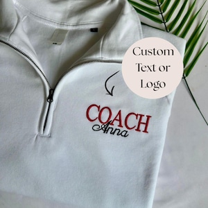 Op de afbeelding: Een wit sweatshirt met een korte rits en een borduursel op de linkerborst. Het borduursel bevat het woord COACH in rode hoofdletters, met daaronder de naam Anna in zwart cursief schrift. Een donkere rits loopt door het midden.