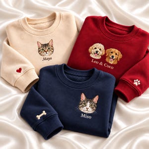 Könnte beinhalten: Drei Rundhals-Sweatshirts in Beige, Dunkelrot und Marineblau. Jedes zeigt gestickte Tierporträts mit den Namen: Maya, Leo & Coco und Miso. Der beige Ärmel hat ein rotes Herz, der marineblaue einen Knochen und der rote einen Pfotenabdruck.