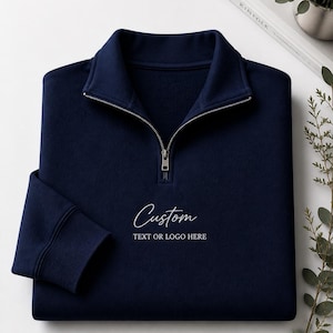 Könnte beinhalten: Marineblaues Sweatshirt mit Viertelreißverschluss und silbernem Reißverschluss. Das Sweatshirt trägt das Wort "Custom" in Schreibschrift, darunter der Text "TEXT OR LOGO HERE". Das Sweatshirt ist sauber auf einer weißen Oberfläche gefaltet.