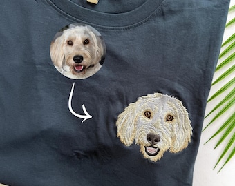 Può includere: Una t-shirt grigio scuro con una fotografia circolare di un cane dal pelo chiaro con orecchie pendenti e bocca aperta. Sotto la foto, una freccia bianca indica un ricamo dettagliato del muso del cane sul petto della t-shirt.