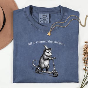 May include: A blue Comfort Colors t-shirt with the phrase "Off to commit Shenanigans" and a graphic of an opossum riding a scooter. A gold star pendant necklace is draped across the shirt.