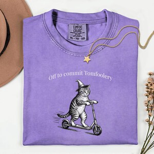 Może przedstawiać: Liliowa koszulka Comfort Colors z napisem "Off to commit Tomfoolery". Koszulka przedstawia grafikę kota w kapeluszu czarownicy jadącego na skuterze. Złoty naszyjnik z wisiorkiem w kształcie gwiazdy jest zarzucony na koszulkę.