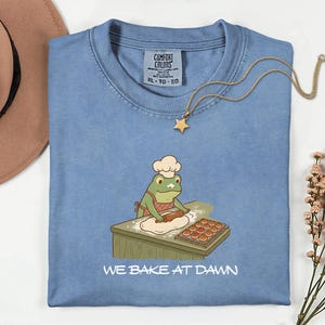 May include: A light blue t-shirt with a frog chef graphic rolling dough, a gold star necklace, and the text "WE BAKE AT DAWN". The shirt has a crew neck and a tag that says "Comfort Colors".