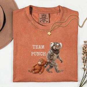 Puede incluir: Camiseta naranja quemado con el texto "TEAM PUNCH" y un gráfico de dos monos. Un mono está sentado y el otro caminando. Un collar con colgante de estrella dorada está sobre la camiseta.