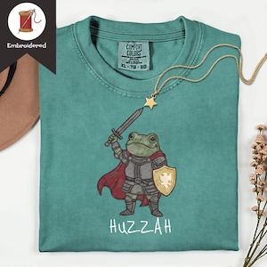 May include: Teal Comfort Colors t-shirt with an embroidered cartoon frog in knight's armour, holding a sword and shield. The word "Huzzah" is printed below the frog. A gold star pendant necklace is draped on the shirt.