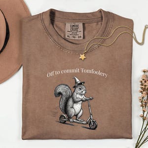 May include: A brown Comfort Colors t-shirt featuring the text "Off to commit Tomfoolery" and a graphic of a squirrel on a scooter wearing a witch's hat. A gold star pendant necklace is on the shirt.