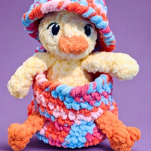 Könnte beinhalten: Ein handgefertigtes, plüschiges Entenküken-Spielzeug mit gelbem Körper, orangefarbenem Schnabel und Füßen sowie schwarzen Augen. Es trägt einen bunten gehäkelten Hut und eine passende Wickel in Blau-, Rosa- und Orangetönen. Das Spielzeug ist vor einem lila Hintergrund platziert.