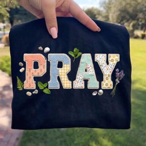 Könnte beinhalten: Schwarzes T-Shirt mit dem Wort "PRAY" aus bunten Stoffen. Die Buchstaben sind mit floralen, Ananas- und Vichy-Mustern verziert. Kleine Steine, Minzzweige und Lavendelzweige sind um die Buchstaben angeordnet.