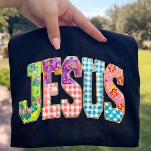 Peut inclure: T-shirt noir avec le mot "JESUS" en lettres brodées colorées. Les lettres présentent des motifs floraux, vichy et à pois dans des tons de vert, violet, orange, rose et bleu. Les lettres sont soulignées en or.
