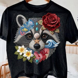 May include: Black t-shirt featuring a raccoon face with floral and ocean-themed embellishments. The raccoon has a red rose, starfish, and seashell accents. The design includes a jellyfish, fish, and coral.