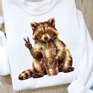 Könnte beinhalten: Weißes Sweatshirt mit einem Waschbär-Motiv, der eine Sonnenbrille trägt und ein Getränk hält. Der Waschbär ist mit braunen und goldenen Pailletten verziert. Der Waschbär zeigt mit seiner Pfote das Friedenszeichen.