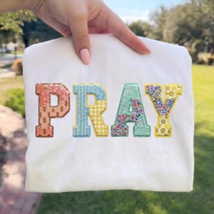 May include: White t-shirt featuring the word "PRAY" embroidered in a variety of colours and textures. The "P" is peach, the "R" is blue and yellow checked, the "A" is floral, and the "Y" is yellow with a floral design.