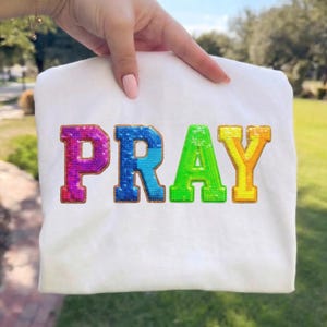 Puede incluir: Camiseta blanca con la palabra "PRAY" en letras de lentejuelas de colores. Las letras van del morado al azul, verde y amarillo. La camiseta está sujeta por una mano con esmalte de uñas rosa, con un fondo de hierba verde.