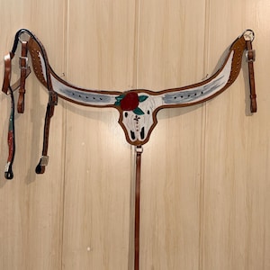 Puede incluir: Un petral de cuero color canela para caballo con un dise&ntilde;o central de cr&aacute;neo de toro blanco, una rosa roja y hojas verdes. La pieza incluye hebillas, remaches y correas ajustables de metal plateado, con detalles pintados en blanco en el yugo.