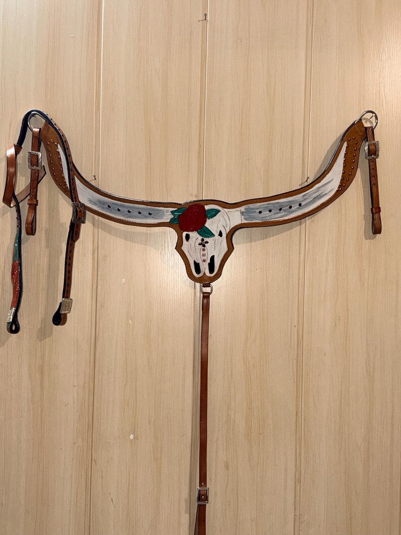 Puede incluir: Un petral de cuero color canela para caballo con un dise&ntilde;o central de cr&aacute;neo de toro blanco, una rosa roja y hojas verdes. La pieza incluye hebillas, remaches y correas ajustables de metal plateado, con detalles pintados en blanco en el yugo.