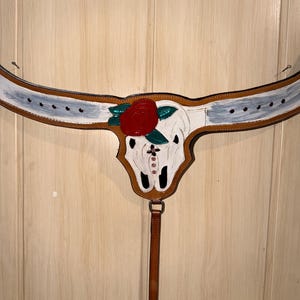 Puede incluir: Un pretal de cuero color canela para caballo con un cr&aacute;neo de toro pintado de blanco en el centro. El cr&aacute;neo est&aacute; adornado con una gran rosa roja, hojas verdes y peque&ntilde;os diamantes de imitaci&oacute;n rosas. Las correas horizontales tienen detalles blancos.