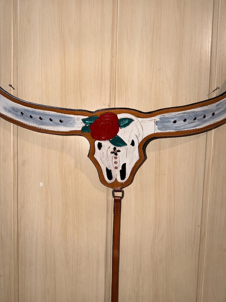 Puede incluir: Un pretal de cuero color canela para caballo con un cr&aacute;neo de toro pintado de blanco en el centro. El cr&aacute;neo est&aacute; adornado con una gran rosa roja, hojas verdes y peque&ntilde;os diamantes de imitaci&oacute;n rosas. Las correas horizontales tienen detalles blancos.