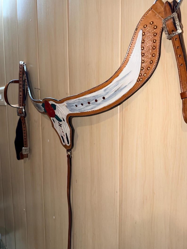Puede incluir: Un petral de cuero color canela para caballo con un dise&ntilde;o pintado a mano de un cr&aacute;neo de toro blanco y una rosa roja. La pieza incluye hebillas de metal plateado, tachuelas decorativas color cobre y correas de cuero ajustables.