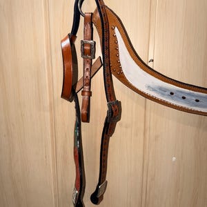 Puede incluir: Una brida de caballo de cuero marr&oacute;n con hebillas de metal plateado y tachuelas decorativas. La testera muestra una secci&oacute;n pintada de blanco con acabado desgastado, mientras que las carrilleras incluyen detalles pintados en rojo y verde.