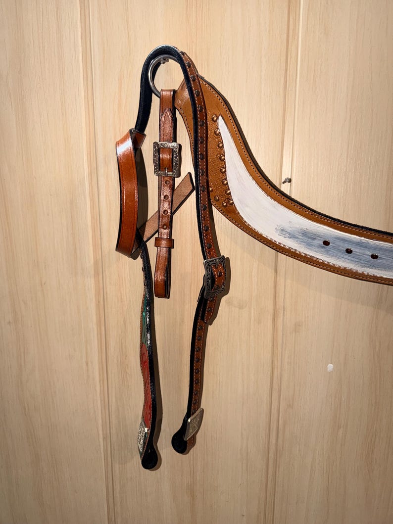 Puede incluir: Una brida de caballo de cuero marr&oacute;n con hebillas de metal plateado y tachuelas decorativas. La testera muestra una secci&oacute;n pintada de blanco con acabado desgastado, mientras que las carrilleras incluyen detalles pintados en rojo y verde.
