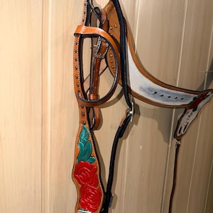 Puede incluir: Una brida de cuero color canela con rosas rojas y hojas verdes pintadas a mano en las carrilleras. La brida incluye herrajes met&aacute;licos plateados, hebillas y conchos cuadrados decorativos, con forro y costuras en color negro.