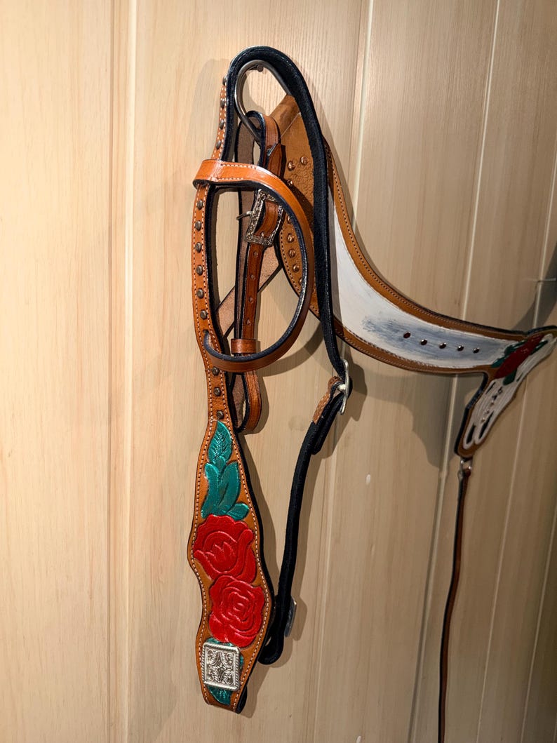 Puede incluir: Una brida de cuero color canela con rosas rojas y hojas verdes pintadas a mano en las carrilleras. La brida incluye herrajes met&aacute;licos plateados, hebillas y conchos cuadrados decorativos, con forro y costuras en color negro.