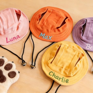 Può includere: Quattro cappelli colorati per animali domestici in rosa, arancione, viola e giallo, ciascuno ricamato con un nome: Luna, Max, Bella e Charlie. I cappelli hanno un sottogola e sono progettati per animali domestici. Un giocattolo a forma di zampa in peluche è in primo piano.