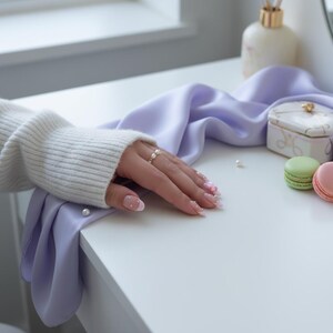 Peut inclure: Une main ornée d'une bague en or avec des perles repose sur un tissu de satin violet clair. Les ongles sont manucurés. Une petite boîte décorative et deux macarons sont posés sur une surface blanche. Un flacon de parfum est en arrière-plan.