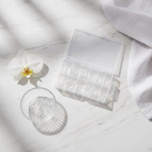 May include: A clear plastic case filled with clear artificial nail tips. A small glass bowl with more nail tips sits next to the case. A white orchid flower and a white towel are also in the image.