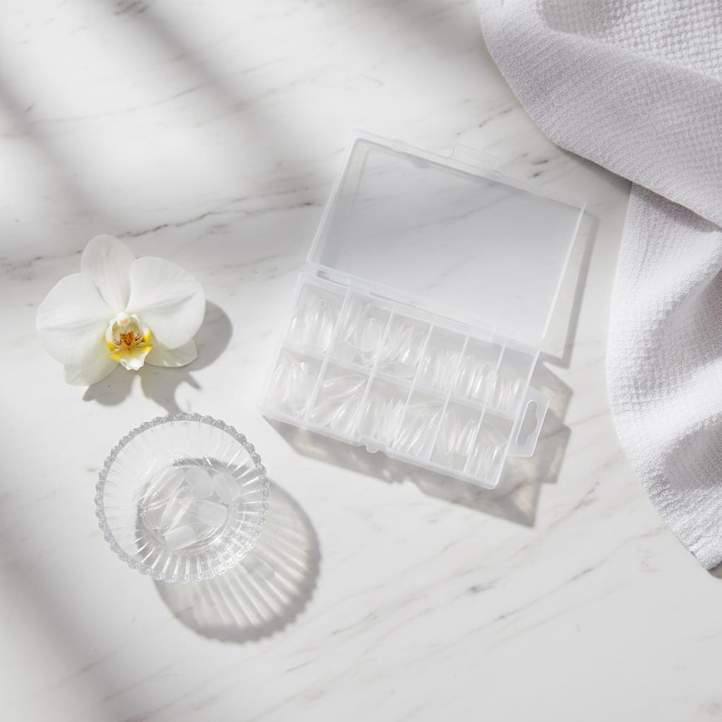 May include: A clear plastic case filled with clear artificial nail tips. A small glass bowl with more nail tips sits next to the case. A white orchid flower and a white towel are also in the image.