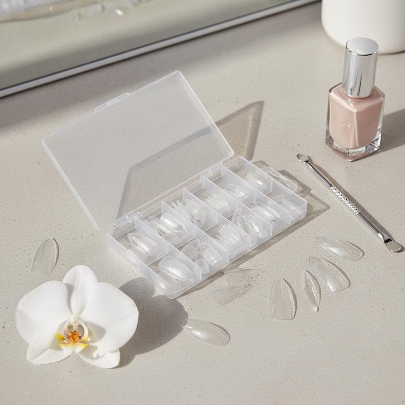 May include: A clear plastic box containing oval-shaped false nails, with several nails scattered around. A bottle of light pink nail polish and a metal cuticle pusher are also present. A white orchid flower is in the foreground.