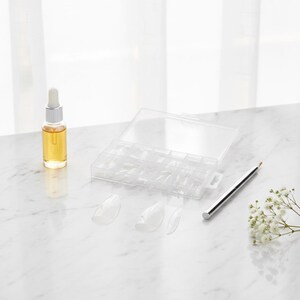 May include: A clear plastic organizer filled with artificial nail tips, a small glass bottle with a dropper, and a silver nail art pen are arranged on a white surface. Three clear nail tips are in the foreground. A sprig of white flowers is in the background.