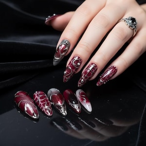 Peut inclure: Ensemble de faux ongles longs et pointus, ornés de motifs complexes. Les ongles présentent un dégradé de rouge foncé à blanc, avec des détails argentés. L'ensemble est présenté sur une surface noire.